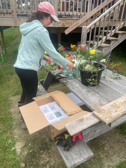 A woman in a pink hat and light blue sweatshirt arranging flowers in a vase on a picnic table