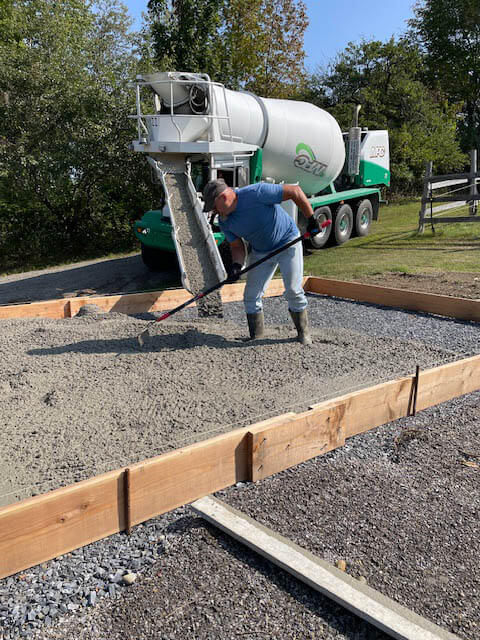 A man spreads wet concrete with a rake on a prepared foundation as a cement mixer truck pours more concrete, surrounded by trees and grass.