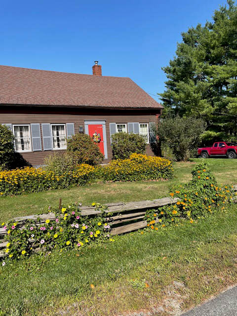 A brick house with a red front door, gray shutters, and yellow flowers in the garden; a red pickup truck is parked on the right side near tall green trees.