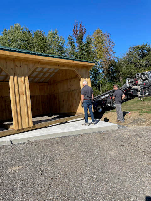 Two people stand next to a newly placed wooden shed on a concrete slab, with a truck and trees in the background under a clear blue sky.